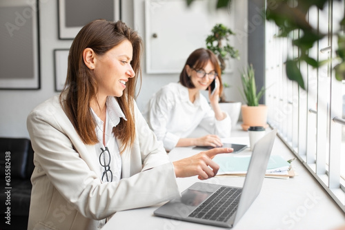 Female company employees working in office lady pointing at laptop screen while colleague talking on phone on background