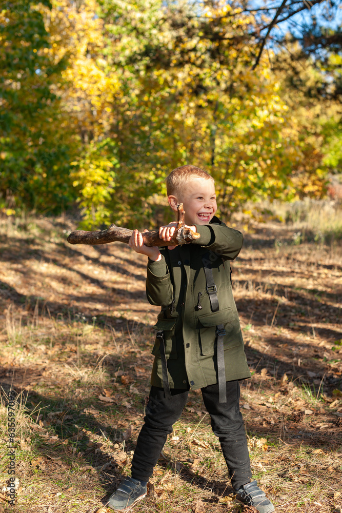 Naklejka premium A little boy in a green jacket plays with a stick in nature in the autumn forest