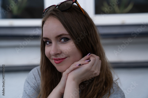 young pretty woman with good skin, beautiful big brown eyes, natural hair color, red lips, sitting in the street and smiling. Girl portrait. Looking at the camera, visible hands © Anna Ivanovska