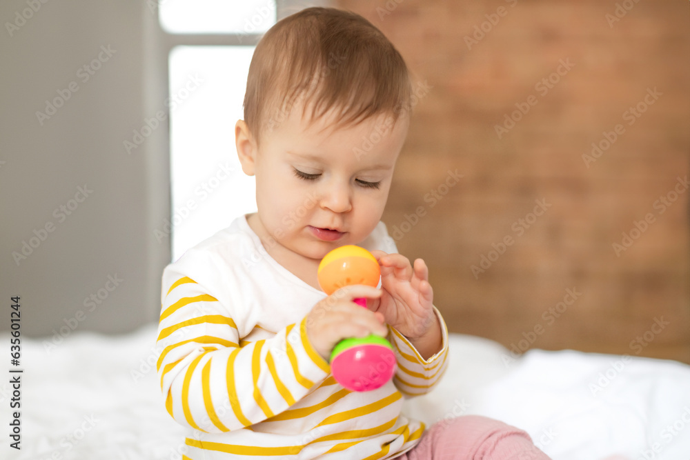 Cute baby girl with rattle toy in hand sitting on bed little child in bodysuit playing, closeup, copy space Cute baby girl with rattle toy in hand sitting on bed little child in bodysuit playing, closeup, copy space