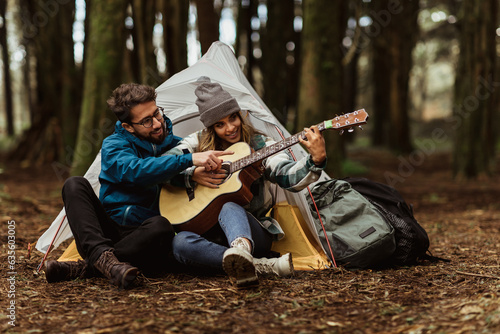 Glad young caucasian guy help lady with music lesson in forest, enjoy travel vacation with tent, playing guitar