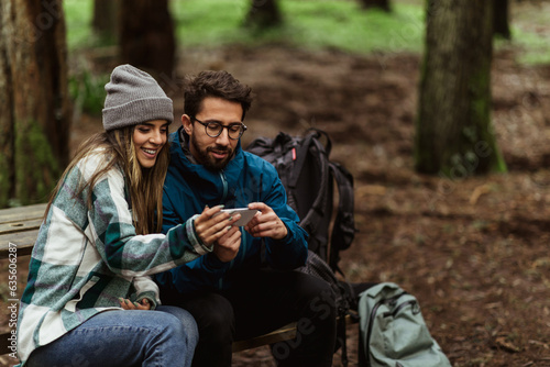 Positive young caucasian couple in jackets in forest enjoy vacation, sit on bench with backpacks, use smartphone app map © Prostock-studio