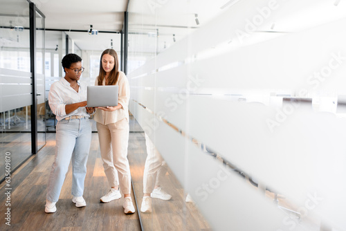 Businesswoman with laptop explaining project details to colleague at office hallway. Business partners having meeting in coworking office space. Two female diverse employees, full length