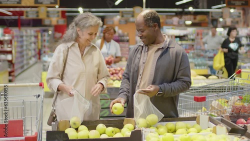 Selective focus medium shot of senior Black man and Caucasian woman choosing one fruit at the same time while picking up apples in supermarket, man giving it to woman