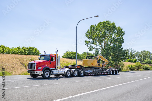 Red day cab powerful big rig semi truck tractor transporting excavator on step down semi trailer driving on the highway road exit
