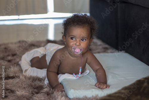 Black baby girl with a pacifier, about 6 months old, smiling and trying to crawl on floor.