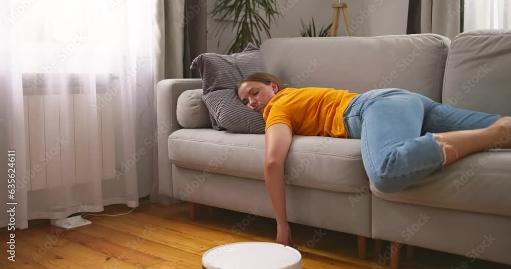 Tired woman sleeping on sofa at home while robot vacuum cleaner working on floor Tired woman sleeping on sofa at home while robot vacuum cleaner working on floor