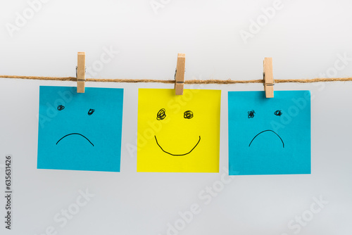 Photos Positive attitude concept, three sticky notes hanging on a string attached with wooden clothespins, two sad faces in blue color and one standing out smiling face in yellow color in the middle