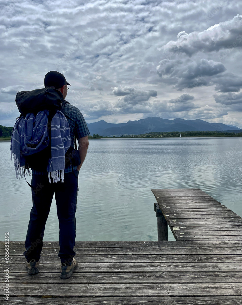 Mann mit Rucksack von hinten steht auf einem Steg am Chiemsee und schaut über den See in die Chiemgauer Alpen, Chiemgau, Bayern, Deutschland