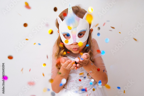 A child in a carnival mask of a unicorn, hand-made from glitter foamiran and faur fux. Little cute cheerful comical girl in the role of a unicorn, preparing for the carnival, masquerade, holidays.