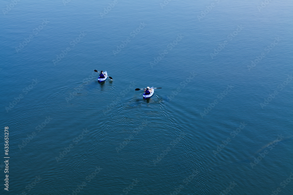 Two Men Paddling Away From Camera On Blue Water With Wake
