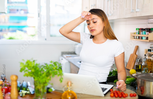 Tired colombian woman looking at laptop during cooking vegetable salad at home