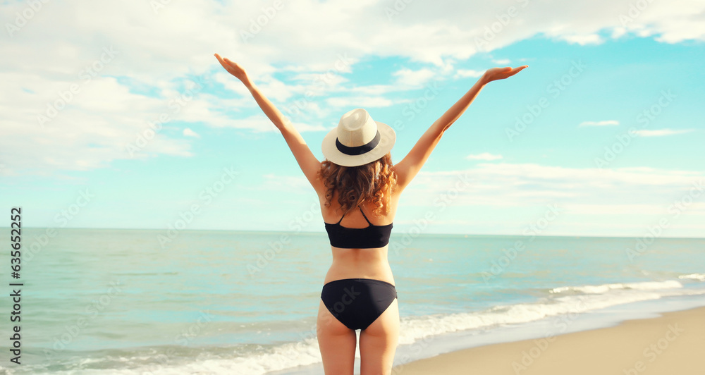 Summer vacation, rear view of beautiful happy slim woman in bikini swimsuit and straw hat raising her hands up on the beach on sunny day on sea coast background Summer vacation, rear view of beautiful happy slim woman in bikini swimsuit and straw hat raising her hands up on the beach on sunny day on sea coast background