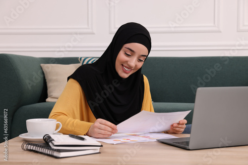 Muslim woman in hijab writing notes near laptop at wooden table indoors