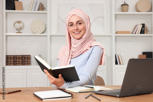 Muslim woman studying near laptop at wooden table in room