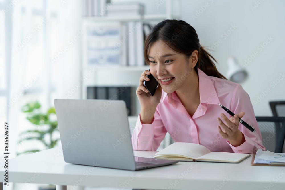 Happy young asian woman talking on the mobile phone and smiling while sitting at her working place in office. Happy young asian woman talking on the mobile phone and smiling while sitting at her working place in office.