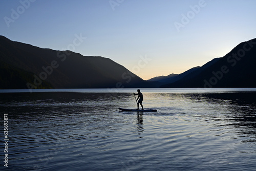 Male paddleboarder in silhouette at sunset on Lake Crescent in Olympic National Park, Washington on calm clear summer afternoon.