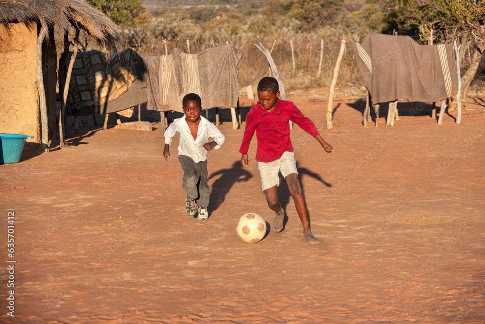village african kids playing soccer on an improvised football field in the yard between the thatched huts homes - obrazy, fototapety, plakaty 