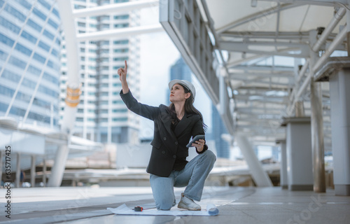 Expert female architect reviews Skytrain site, cross referencing blueprint for project approval. © SpaceOak