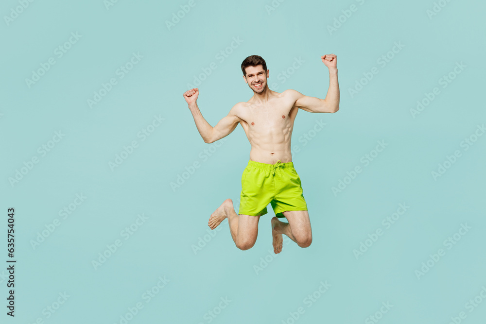 Full body young man wear green shorts swimsuit relax near hotel pool jump high do winner gesture celebrate clenching fists isolated on plain blue background. Summer vacation sea rest sun tan concept.