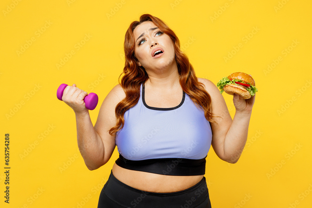 Young tired chubby overweight plus size big fat fit woman wear blue top warm up training hold in hand burger and dumbbells isolated on plain yellow background studio home gym. Workout sport concept. Young tired chubby overweight plus size big fat fit woman wear blue top warm up training hold in hand burger and dumbbells isolated on plain yellow background studio home gym. Workout sport concept.