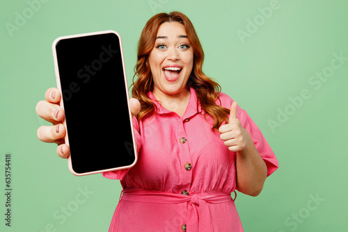 Young fun chubby overweight redhead woman wear casual clothes pink dress hold in hand use close up blank screen workspace area mobile cell phone show thumb up isolated on plain green color background. photo