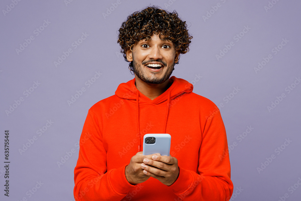 Young surprised smiling Indian man wears red orange hoody casual clothes hold in hand use mobile cell phone isolated on plain pastel light purple color background studio portrait. Lifestyle concept. Young surprised smiling Indian man wears red orange hoody casual clothes hold in hand use mobile cell phone isolated on plain pastel light purple color background studio portrait. Lifestyle concept.