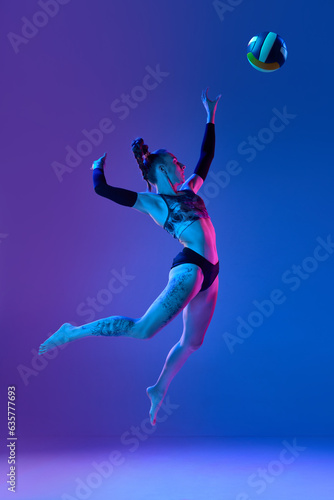 Full-length of young sportive woman, volleyball player in motion, hitting ball in a jump against blue studio background in neon light. Concept of professional sport, competition, health, hobby, ad