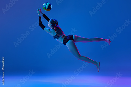 Dynamic image of young woman during game,playing volleyball, hitting ball against blue studio background in neon light. Concept of professional sport, competition, health, hobby, action, ad © master1305