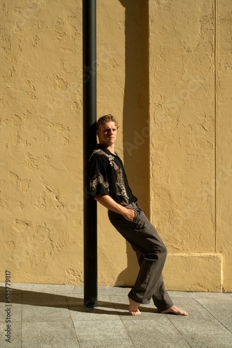 A young blond man wearing a black Hawaiian vintage shirt and gray pants leaning against a lamp post with a yellow wall behind him. © Anissa