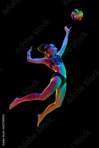 Full-length of sportive young woman, volleyball player in motion, hitting ball in a jump against black studio background in neon light. Concept of professional sport, competition, health, action, ad