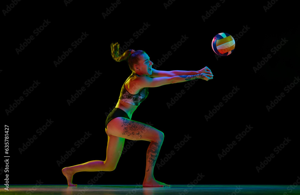 Young competitive woman, volleyball player in motion, training, hitting ball against black studio background in neon light. Concept of professional sport, competition, health, hobby, action, ad