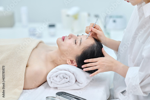 아시아 한국의 젊은 여성이 스파 또는 피부관리전문점 또는 피부과 병원에서 침대에 누워 베이지색 대형 타올을 덮고 눈을 감고있고 하얀색 셔츠를 입은 전문관리사가 여드름 압출기로 문제성피부를 관리하고 있다. 