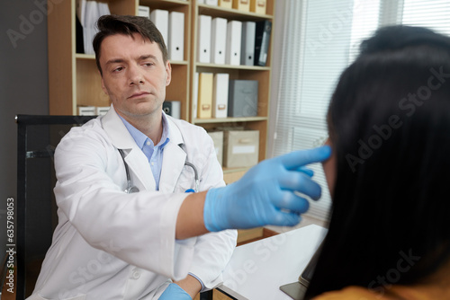 Pensive plastic surgeon in gloves touching face of patient deciding what procedure to recommend