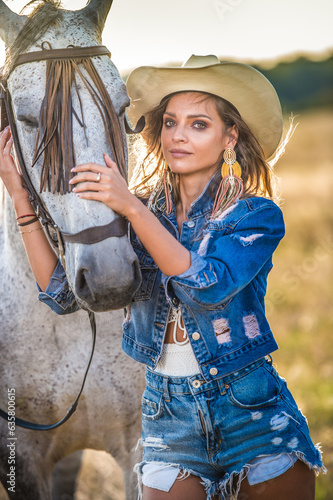 Obraz na płótnie Beautiful blonde woman with curly hair with white hat and horse. Portrait of a girl with denim and her horse. Beautiful girl interacting and having fun with a horse at the ranch w salonie