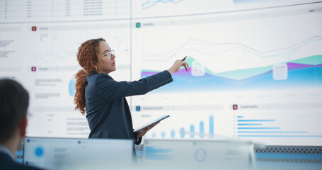Hispanic Female Senior Data Scientist Reviewing Reports Of Risk Management Department On Big Digital Screen In Monitoring Room. Diverse Consulting Company Employees Working Behind Desktop Computers.