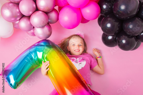 Portrait of a happy little girl lying on a pink background with balloons. and holding the number 7. Happy birthday. Pink background.
