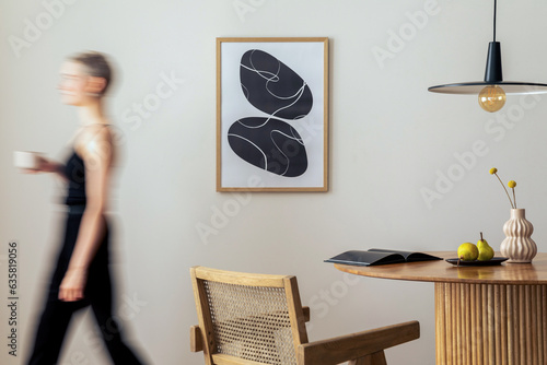 Warm and cozy interior of living room space with mock up poster frame, round table, pedant lamp, rattan chairs and walking young woman. Minimalist home decor. Template. photo