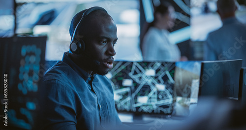 Handsome Surveillance Specialist in a Modern Monitoring Room with Displays and Computers. Black Man Effectively Multitasking with Supporting Clients on a Phone Call and Computer Work