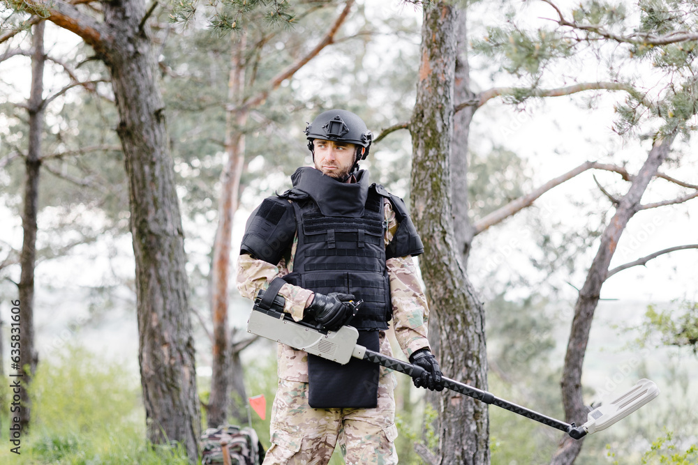 A man in a military uniform and bulletproof vest works in the forest with a metal detector. A minesweeper performs work on demining the territory