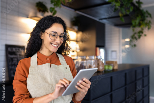 Fotografia Female barista waitress inside cafe using tablet, latin american restaurant owner smiling using tablet app to confirm online order