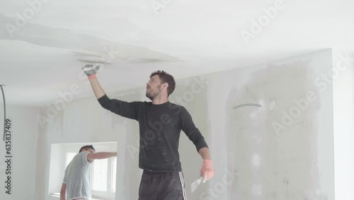 Builders teamwork working indoor in house room plastering ceiling to improve drywalls white surface, room renovation preparation for painting. Construction site. Men at work, caucasian young adults.