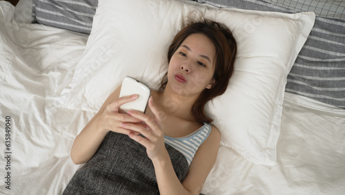 asian female playing with smartphone while lying on bed. careless girl relaxing in bedroom and typing on mobile phone. absent minded woman look at cellphone and fell down hitting her chest in morning