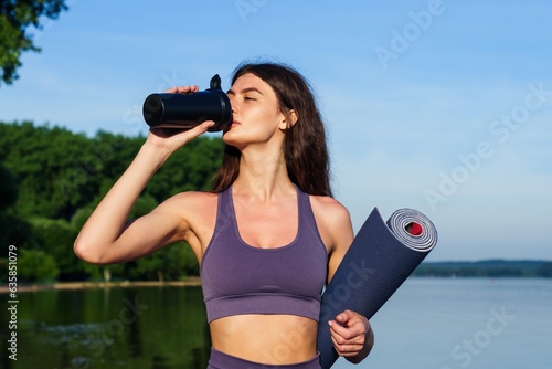 A young woman with a yoga mat outside. An athlete drinks water from a bottle on a summer day outside