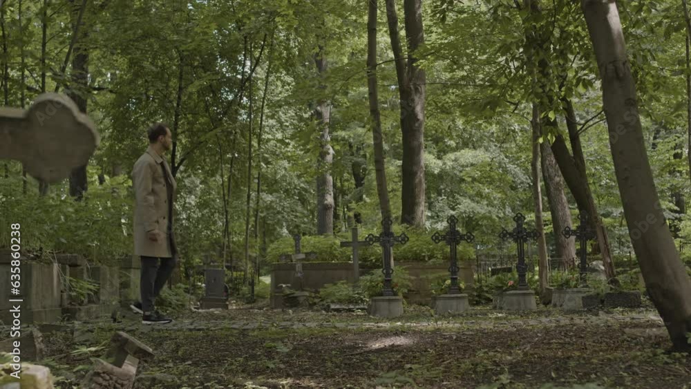 Full length side view slowmo of Caucasian man in camel trench coat walking on graveyard alley at cemetery in daytime Full length side view slowmo of Caucasian man in camel trench coat walking on graveyard alley at cemetery in daytime