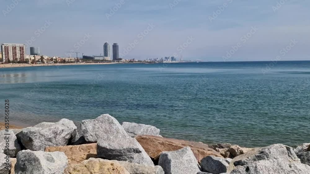 Promenade with stones on the coast of Barcelona in sunny spring. Tall buildings on the horizon near the sandy beach. Promenade with stones on the coast of Barcelona in sunny spring. Tall buildings on the horizon near the sandy beach.