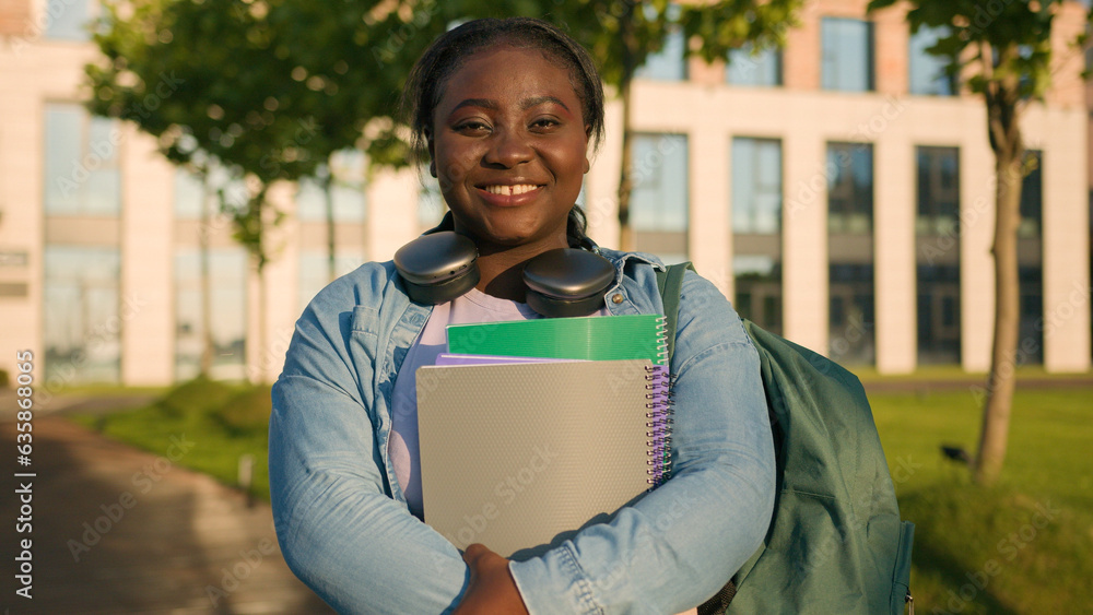 Fototapeta premium Portrait African American woman university college campus academy student teen girl high school pupil holding books with schoolbag headphones smiling toothy joyful in city outdoors study education