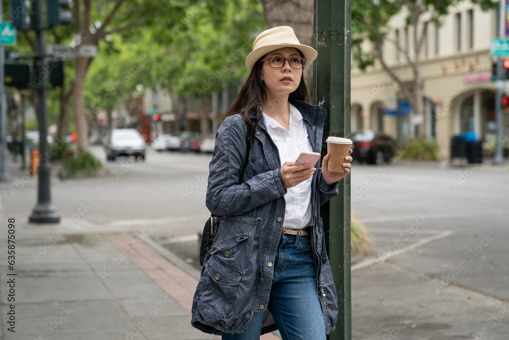 Fototapeta premium asian Japanese female visitor wearing hat and carrying coffee using navigation app on phone on the street while touring palo alto city in California usa in spring