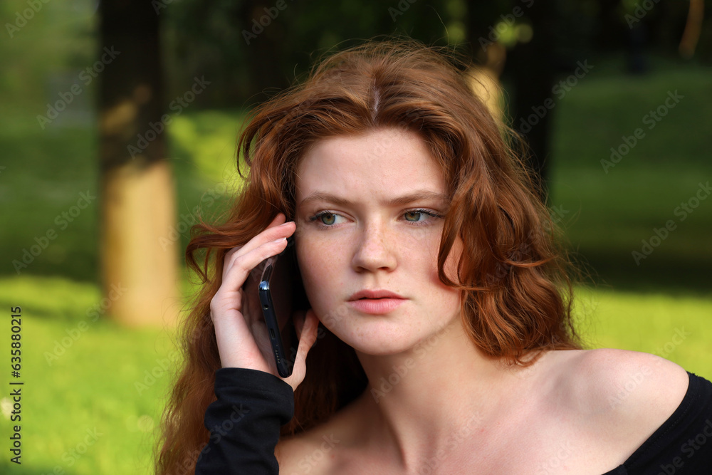 Attractive girl with long red hair and freckles talking on mobile phone sitting on a bench in summer park