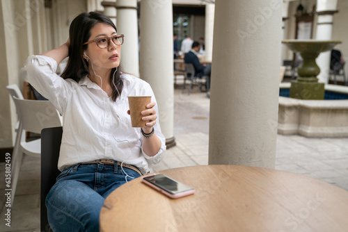asian Japanese businesswoman having coffee and listening to podcast from earphones during break time from work in palo alto city California usa. she looks into space and feels a bit lazy